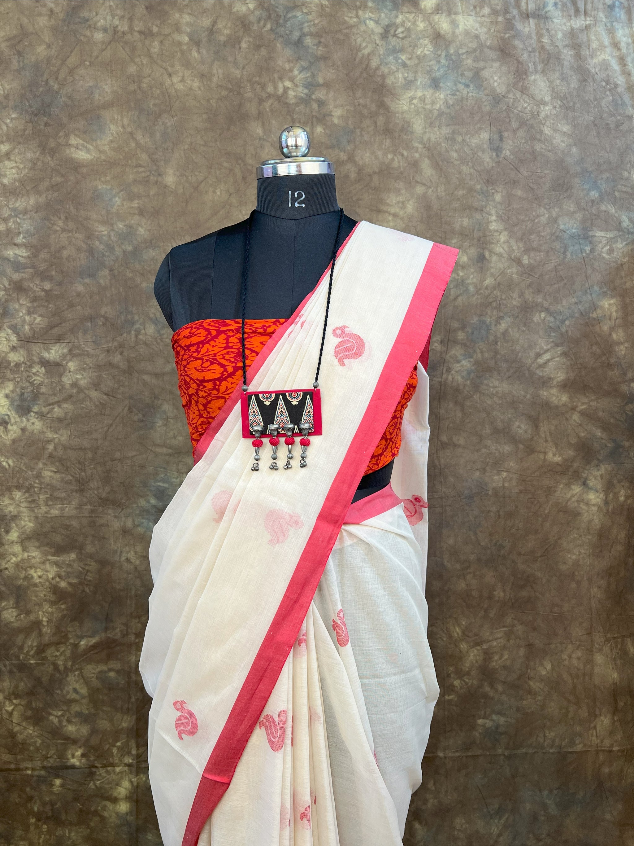 White saree with red border and red blouse on a mannequin against a textured brown background